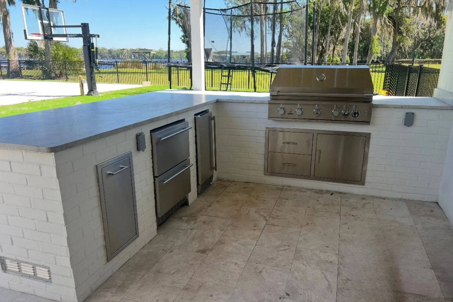 L-shaped outdoor kitchen with stainless steel grill, refrigerator drawers, and travertine flooring by Jax Pavers in Jacksonville FL